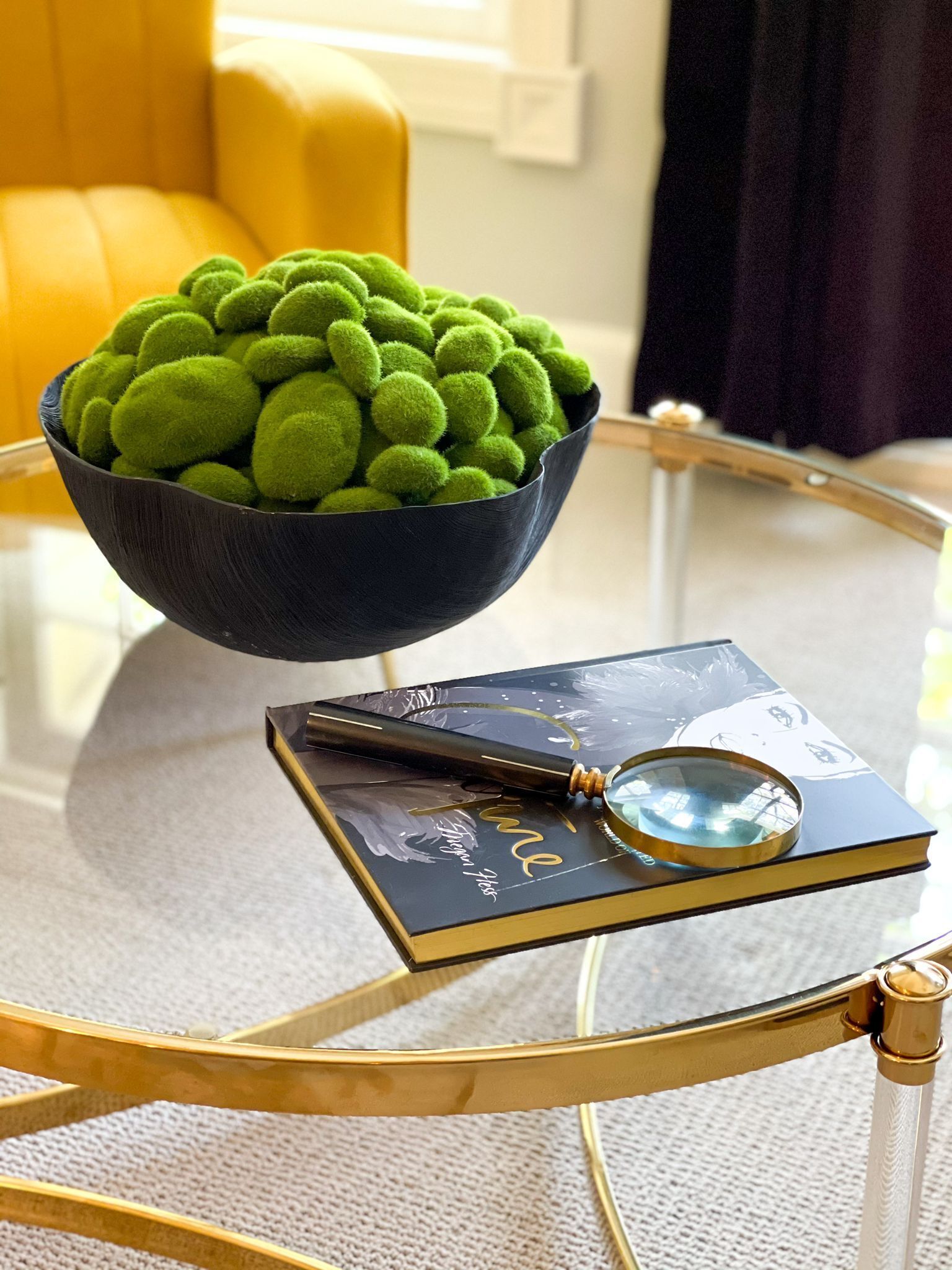 A black bowl with green spherical objects on a glass table, a book, and a magnifying glass.