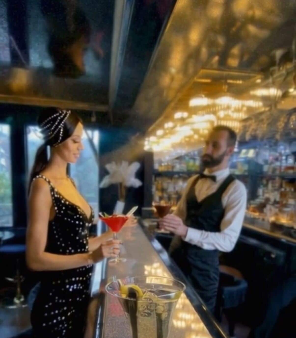 A bar with a bartender and a customer. The bar has a glass counter and shelves with bottles.