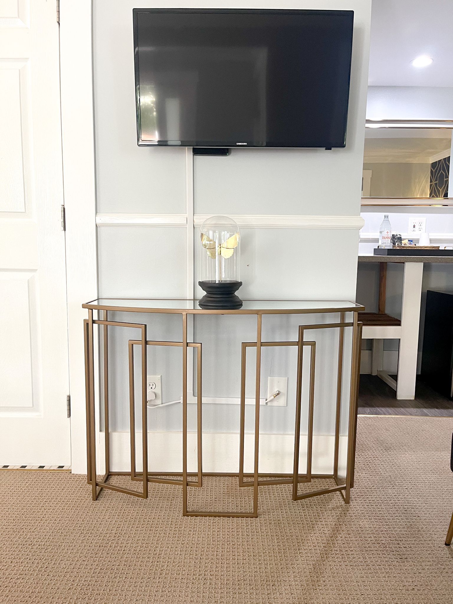 Una sala de estar moderna con un televisor de pantalla plana montado en la pared, una mesa de centro con cubierta de vidrio y un objeto decorativo, y un mueble consola de madera con diseño geométrico.