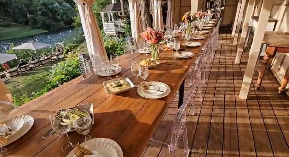 Une longue table en bois, dressée pour un repas avec des assiettes, des verres et des couverts, donnant sur un jardin et un lac.