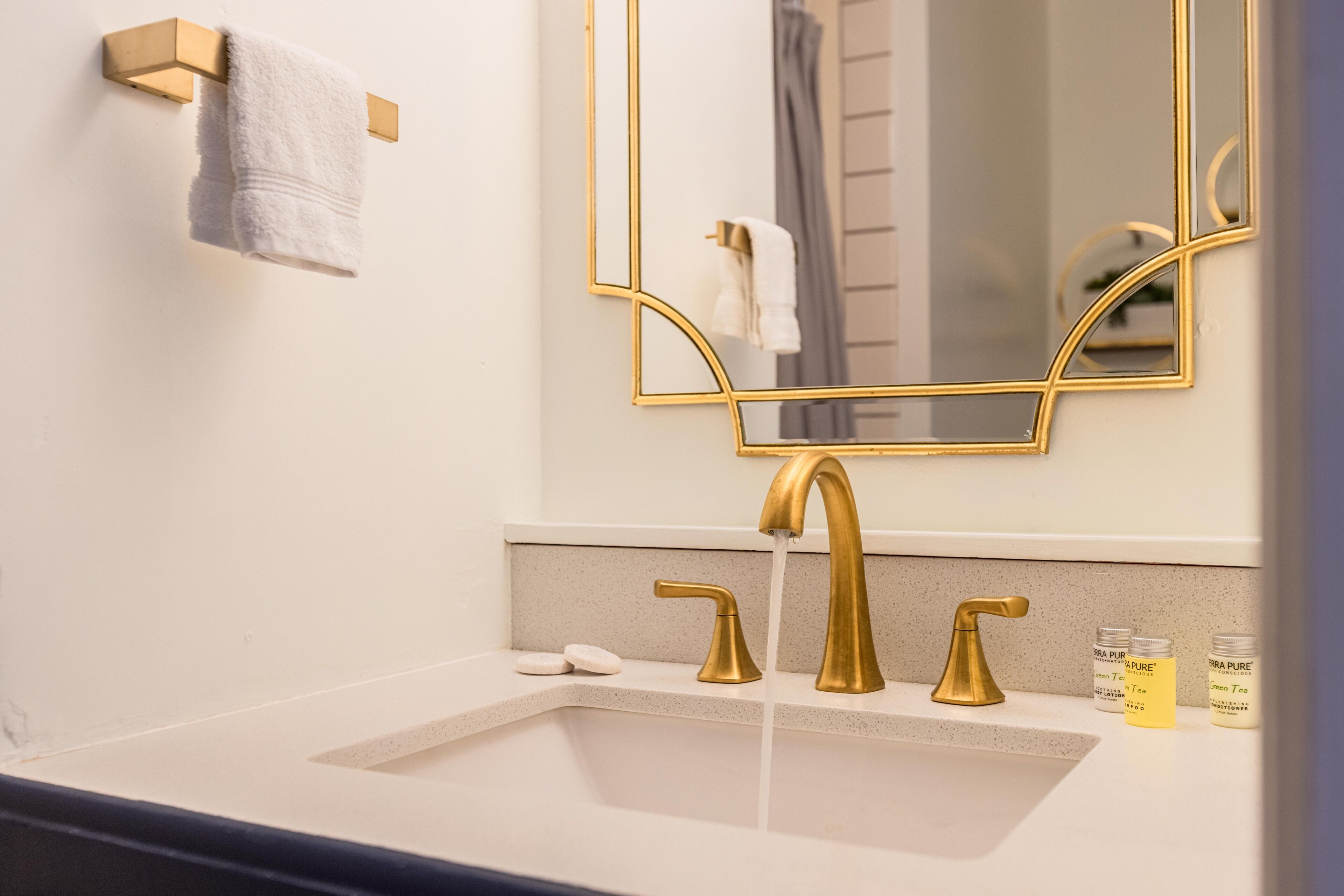 A bathroom with a white sink, gold faucets, and a gold-framed mirror. There are two bottles of green tea on the counter.