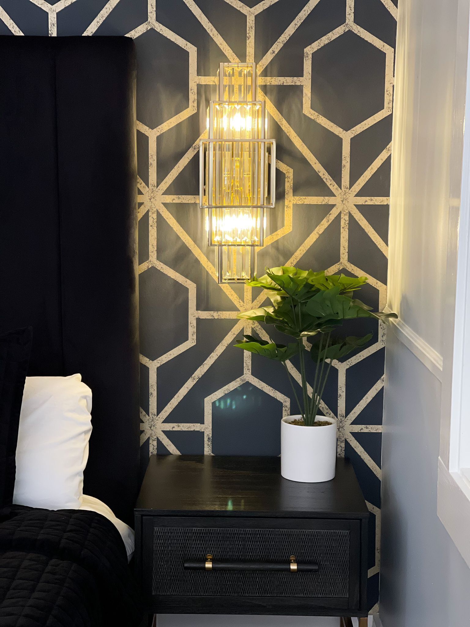 A bedroom with a geometric-patterned wall, a bedside table with a potted plant, and a modern light fixture.