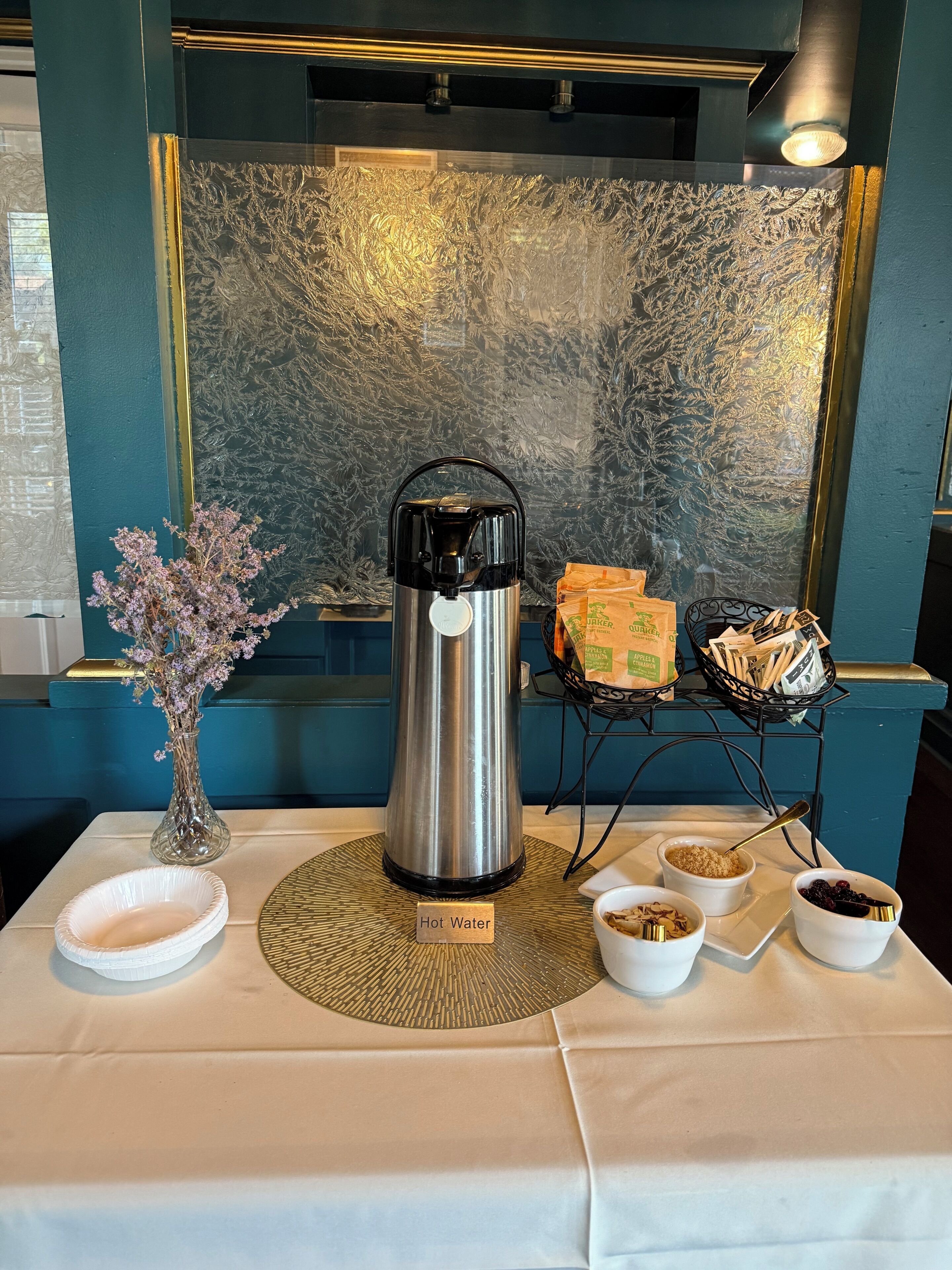 Una mesa con un dispensador de agua caliente, un florero con flores y una bandeja con alimentos.
