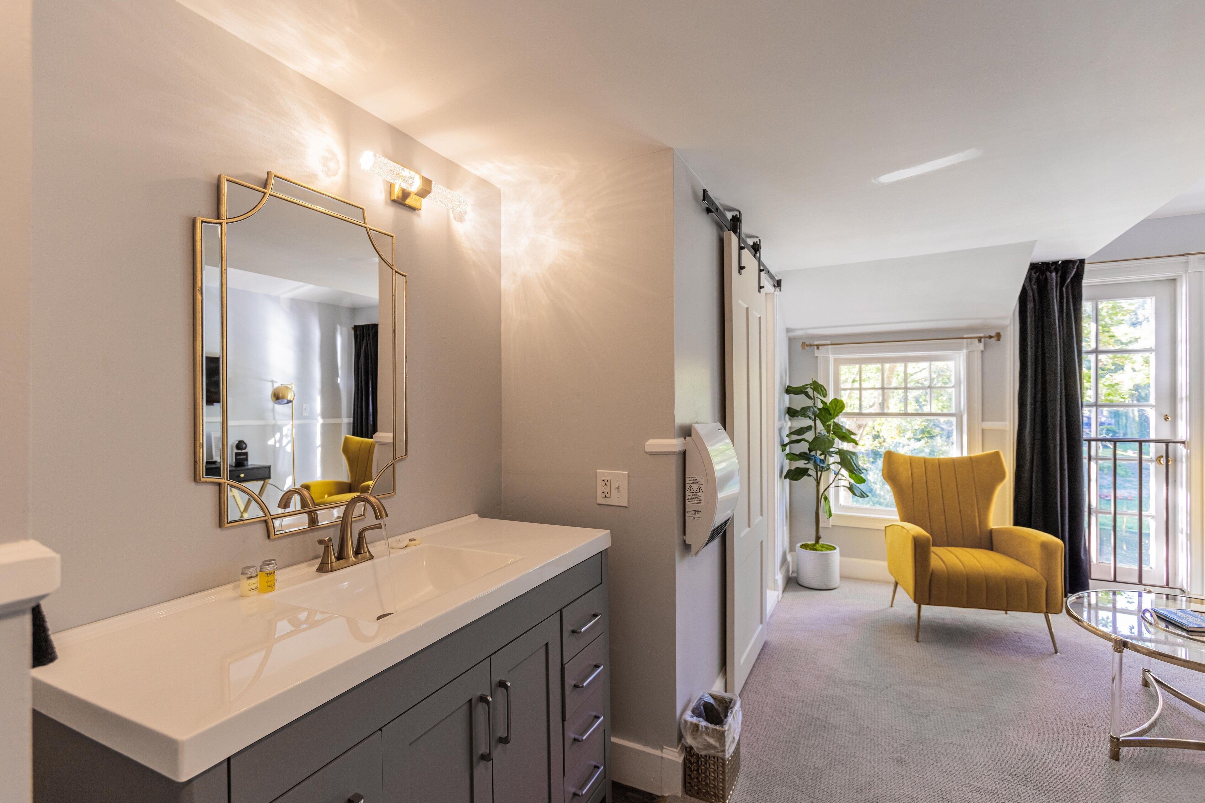 Un baño moderno con un espejo grande, un lavabo blanco y un perchero. Hay una silla y una mesa en la habitación contigua.