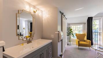 Un baño moderno con un espejo grande, un lavabo blanco y un perchero. Hay una silla y una mesa en la habitación contigua.