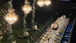 An outdoor dining area with a long table set for a formal event, surrounded by string lights and decorative arches.