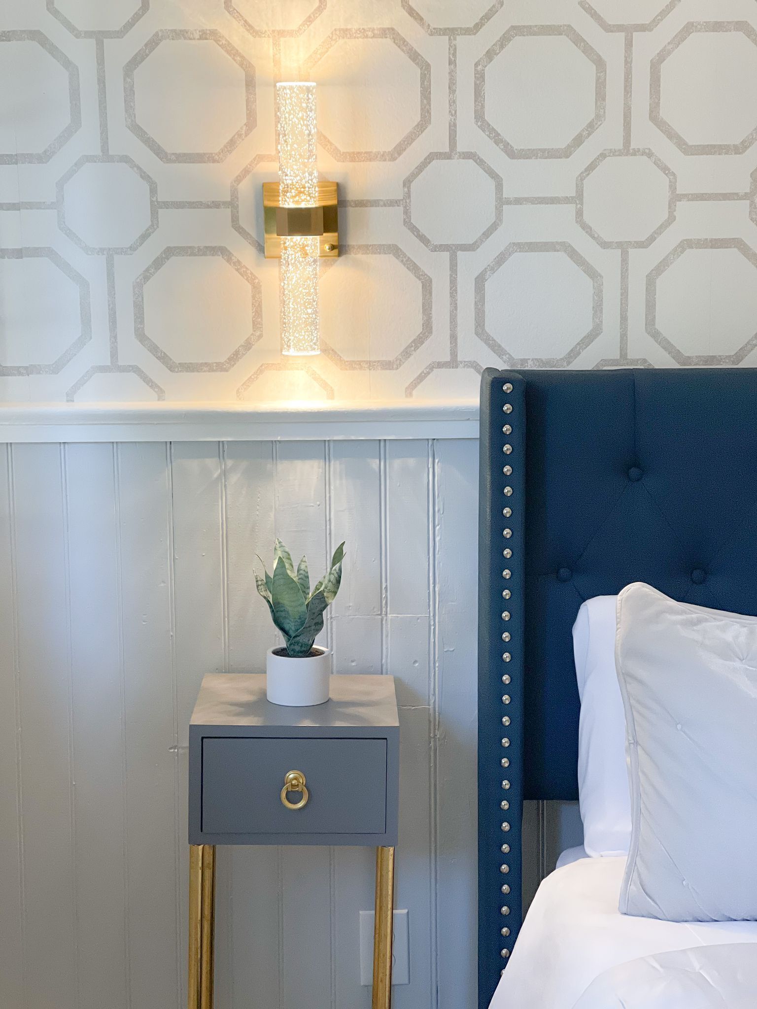 A bedroom with a blue upholstered headboard, a bedside table with a plant, and a wall-mounted light fixture.