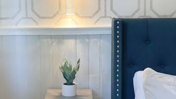 A bedroom with a blue upholstered headboard, a bedside table with a plant, and a wall-mounted light fixture.