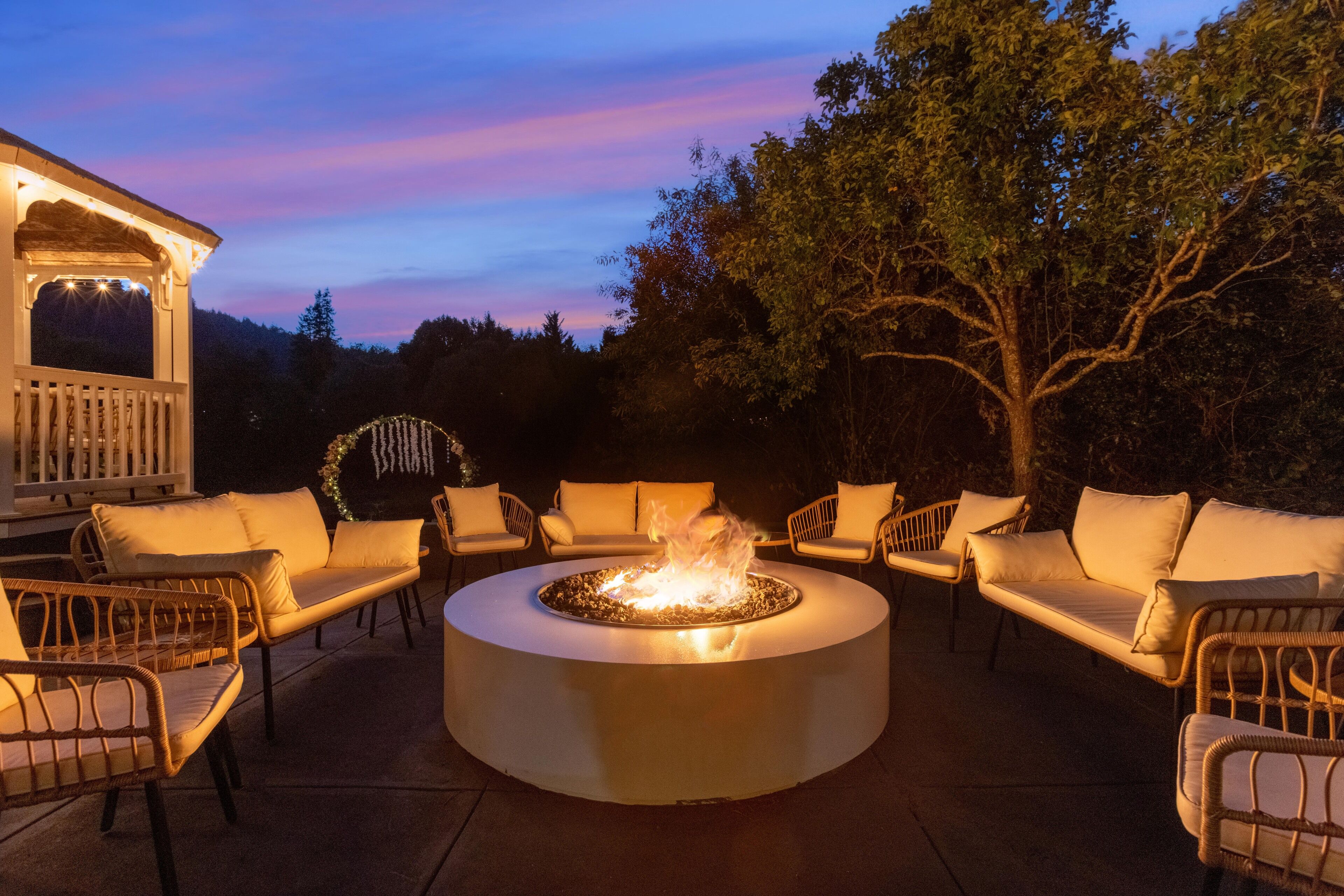 A circular outdoor seating area with a fire feature, surrounded by wicker chairs and a white structure in the background.