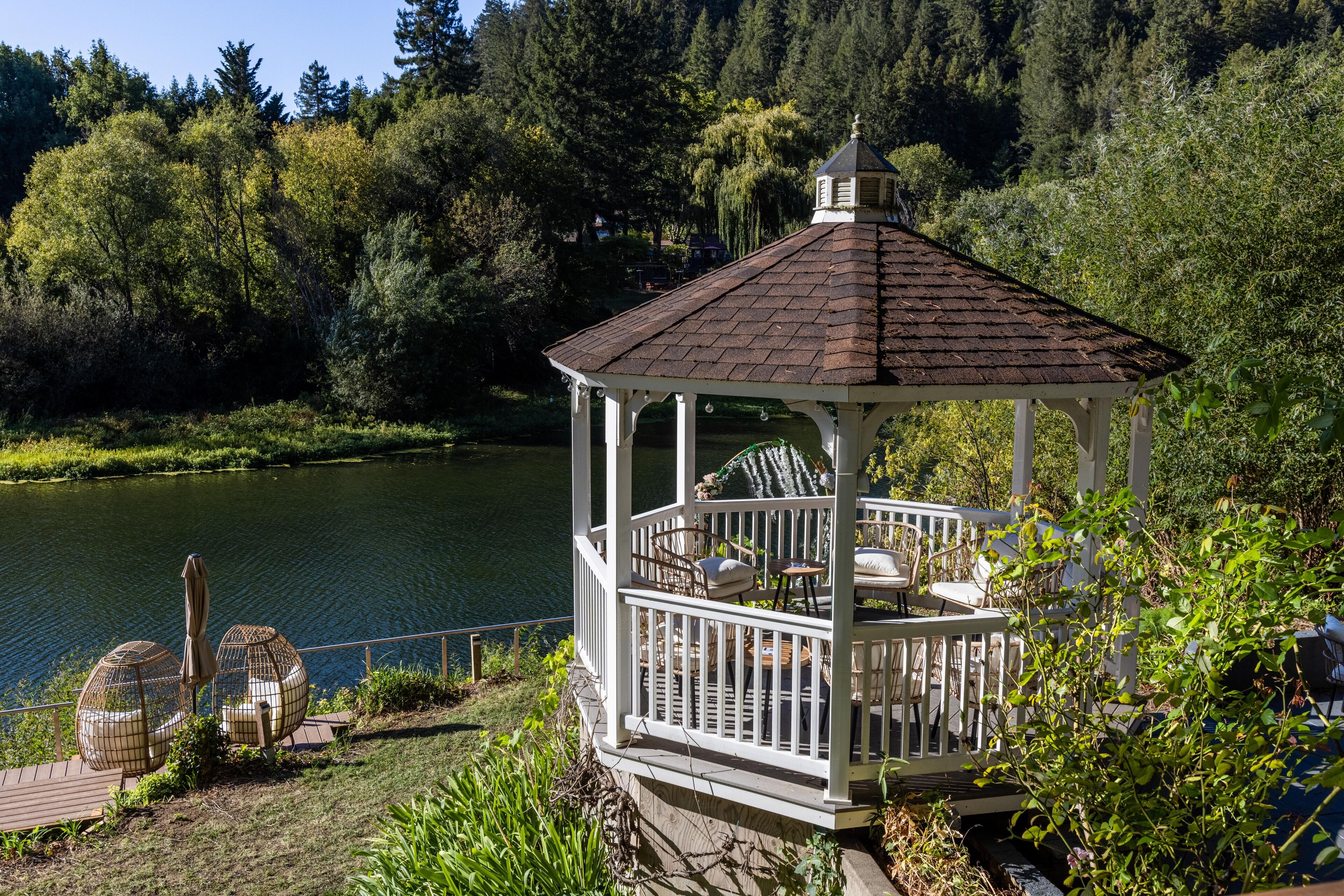 A gazebo with a tiled roof, surrounded by lush greenery and a lake.