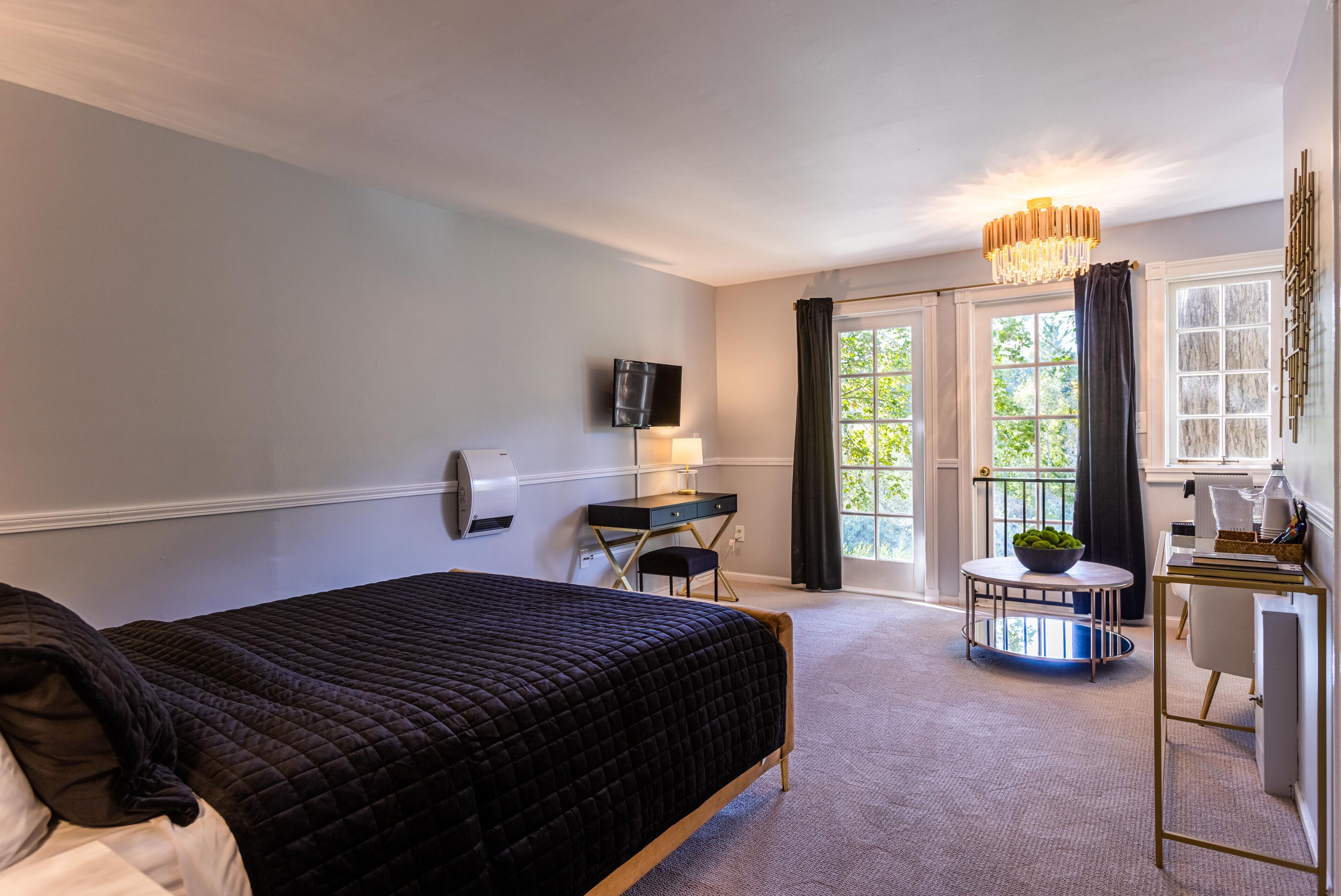 A hotel room with a bed, a desk, a chair, a small table with a bowl, and a chandelier.