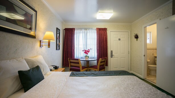 A hotel room with a bed, a desk with a chair, a window with curtains, and a door leading to a bathroom. - Travelers Inn (South San Francisco)