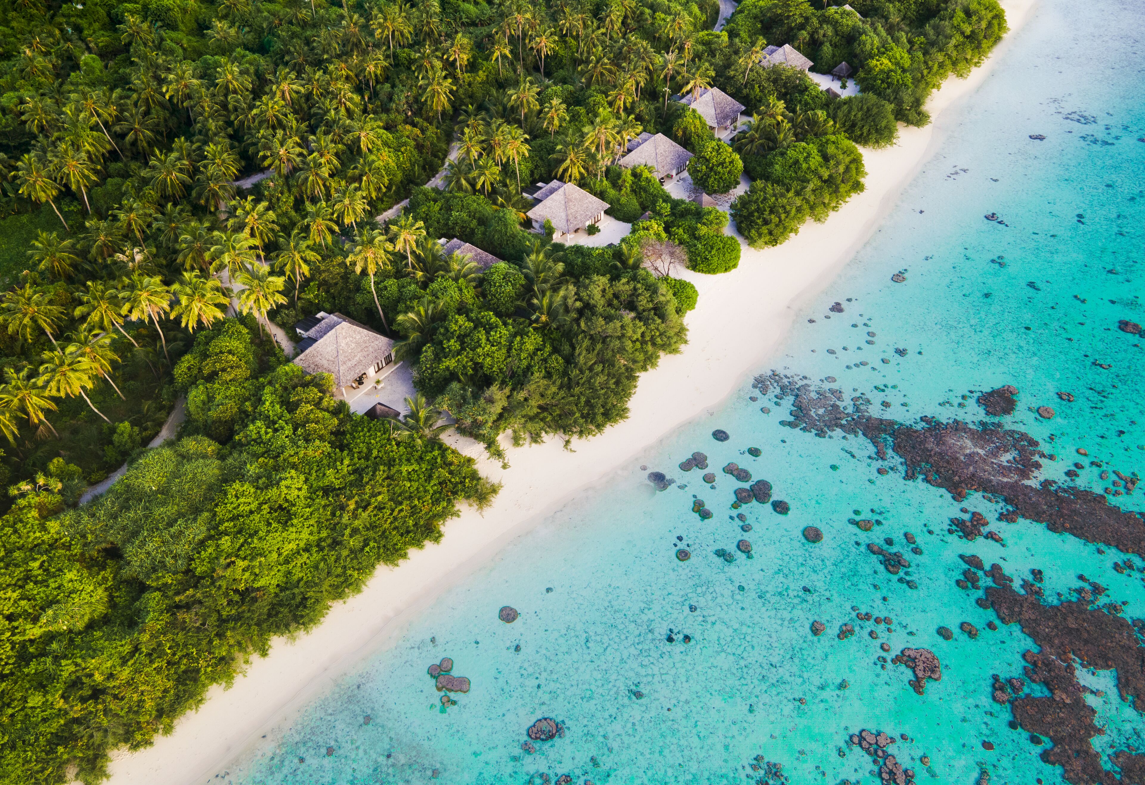 Vista aérea de un resort tropical con bungalows sobre el agua, una playa de arena y aguas turquesas cristalinas.