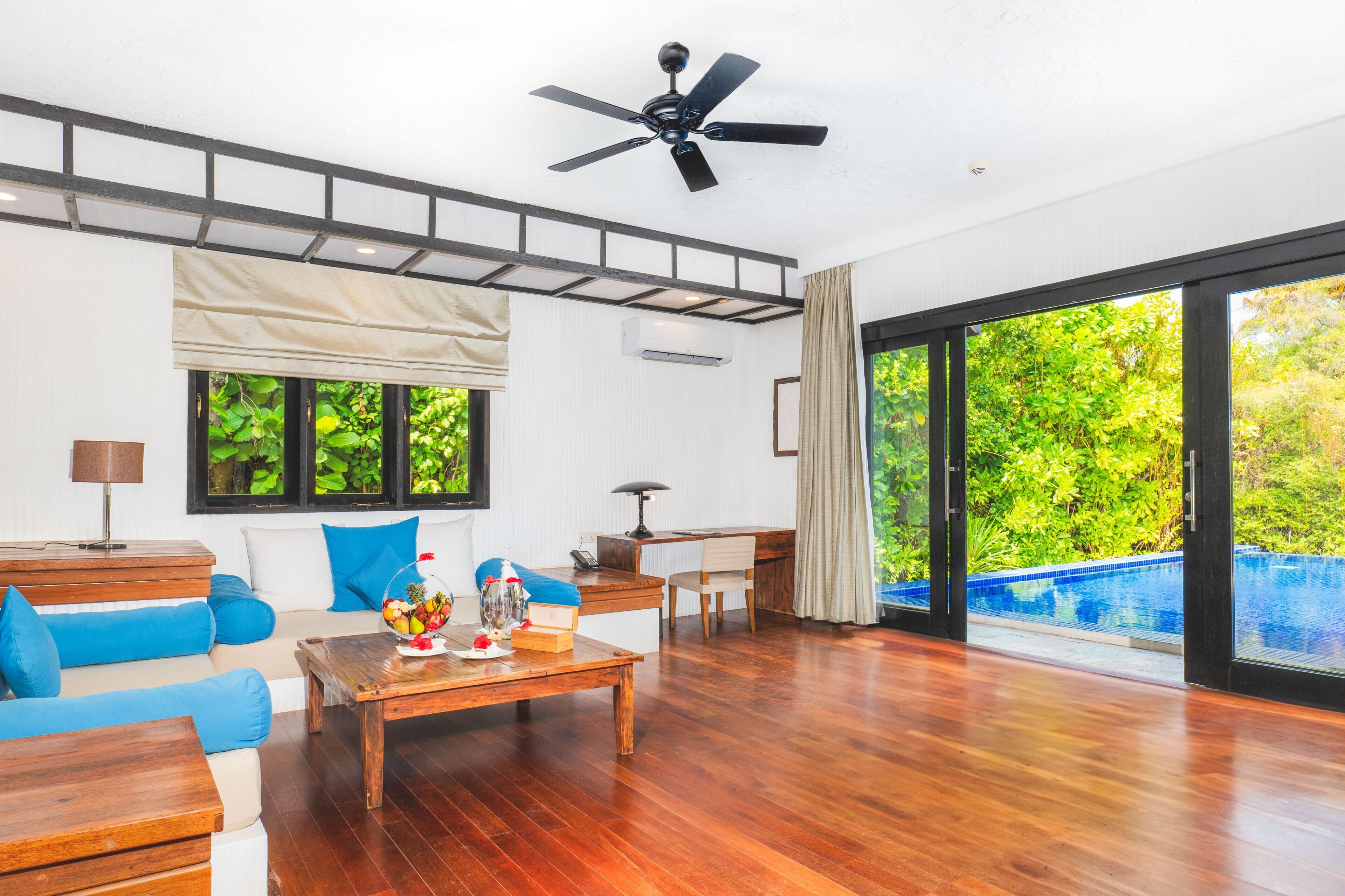 A spacious living area with a wooden floor, a ceiling fan, a sofa with blue cushions, a coffee table with decorative items, a desk, and a view of a swimming pool through large glass doors.