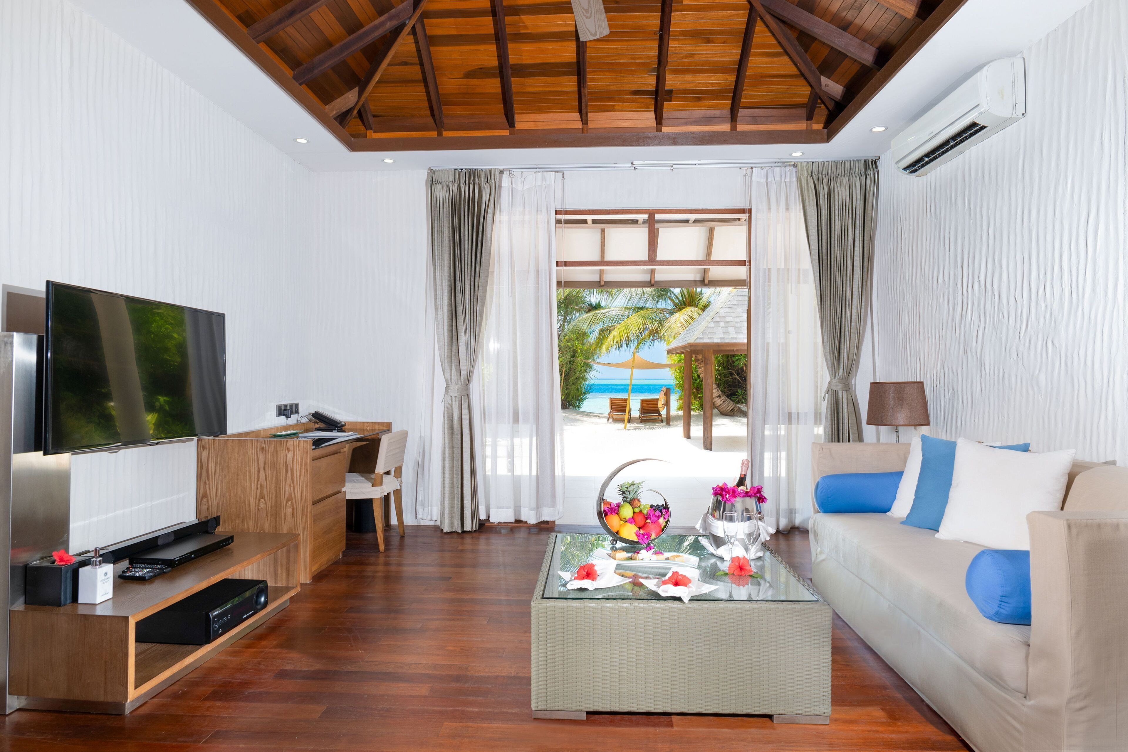 A modern living room with a TV, wooden furniture, a glass coffee table, and a view of a pool and palm trees.