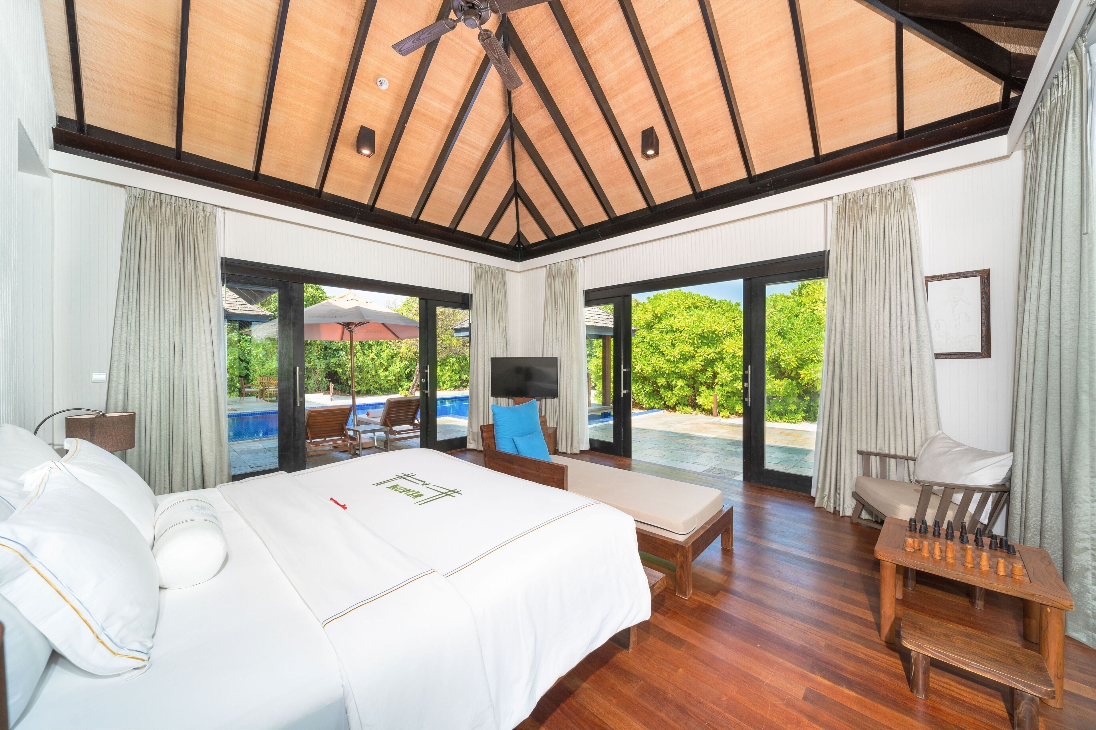 A spacious bedroom with a large bed, a wooden ceiling, and a view of a swimming pool and greenery outside.