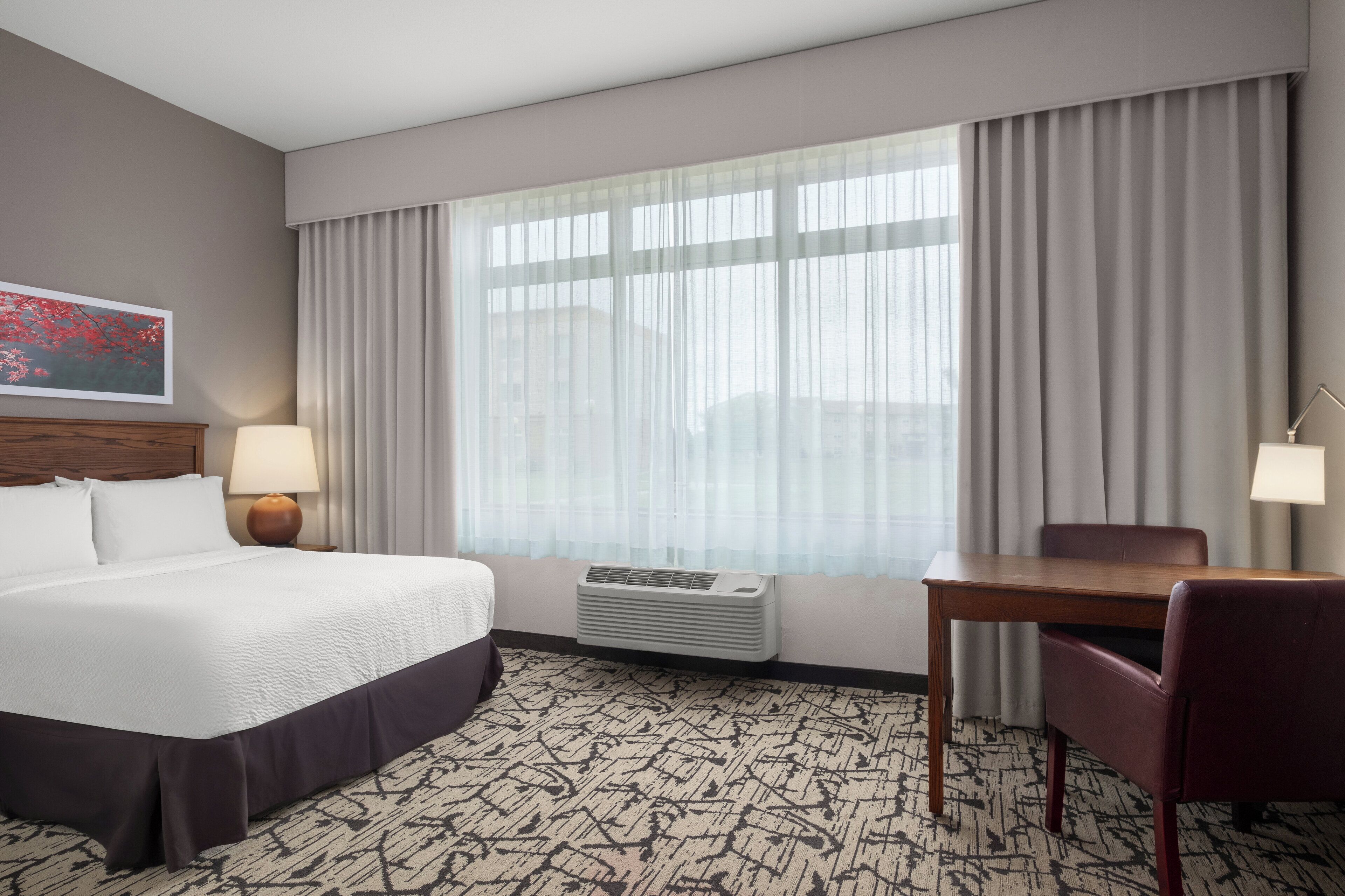 A hotel room with a large bed, a desk, a chair, a lamp, an air conditioner, and a window with curtains.