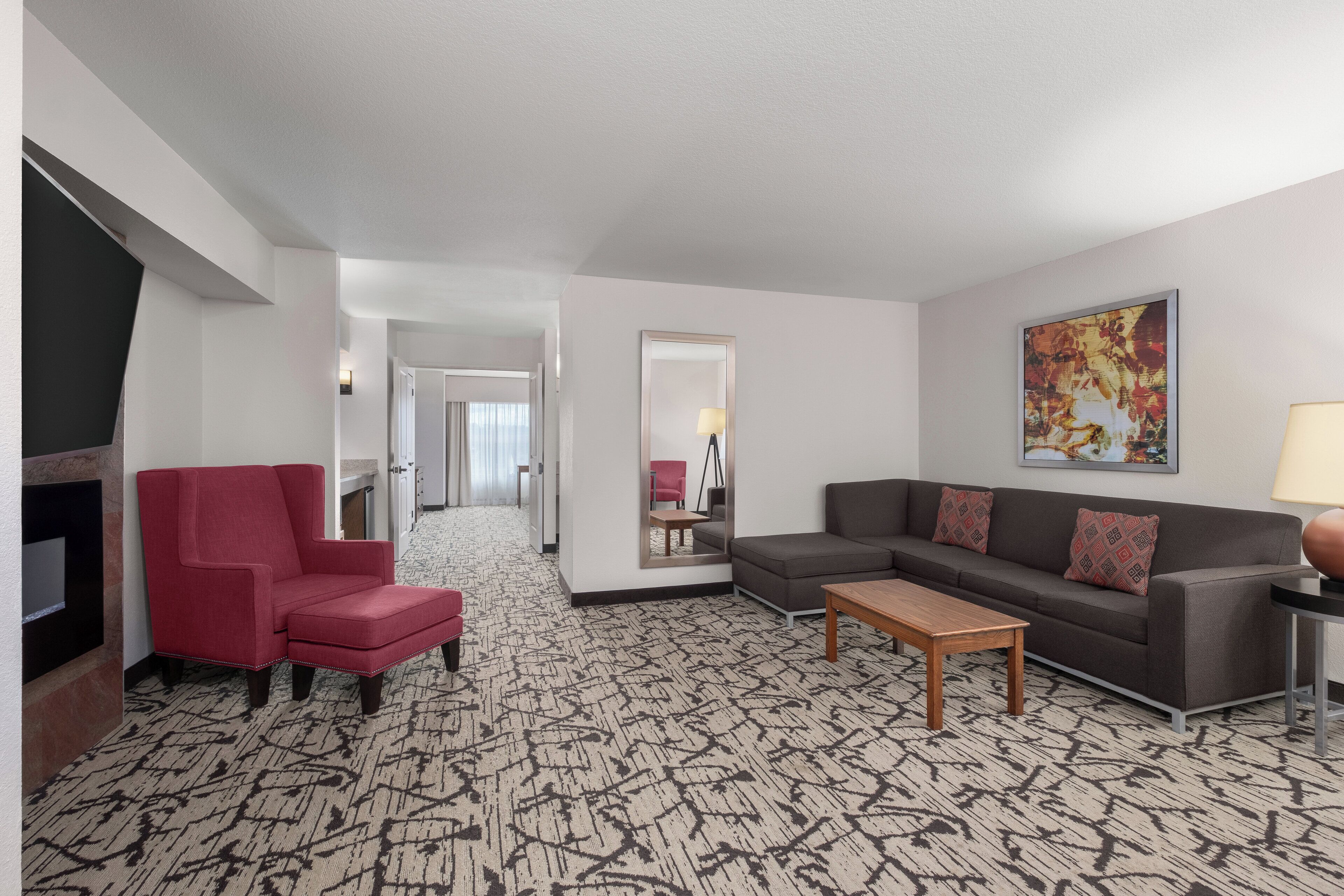 Habitación de hotel con alfombra estampada, un sillón rojo, un sofá gris, una mesa de centro de madera y un cuadro en la pared.