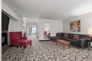 Habitación de hotel con alfombra estampada, un sillón rojo, un sofá gris, una mesa de centro de madera y un cuadro en la pared.
