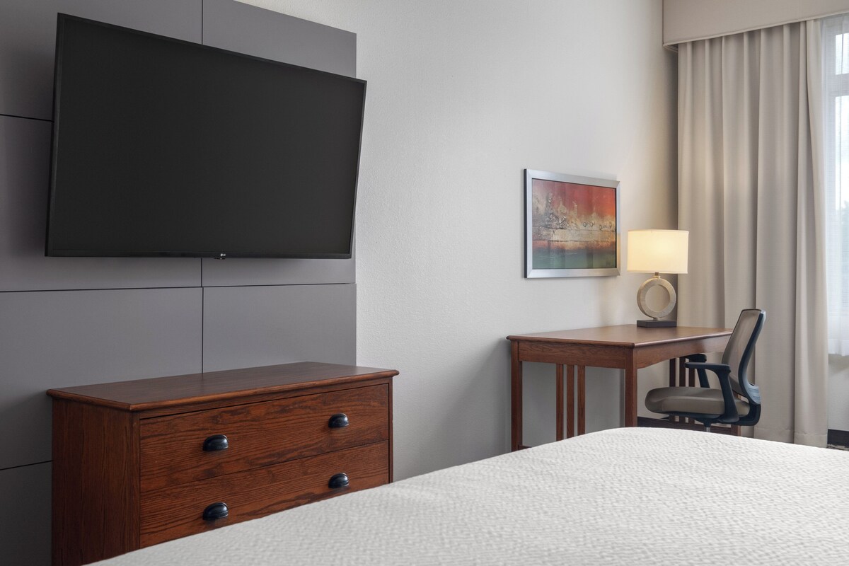 A hotel room with a bed, a wooden dresser, a desk with a chair, a flat-screen TV mounted on the wall, and a painting on the wall.