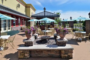Un área de patio exterior con mesas y sillas, rodeada de plantas en macetas y sombrillas.