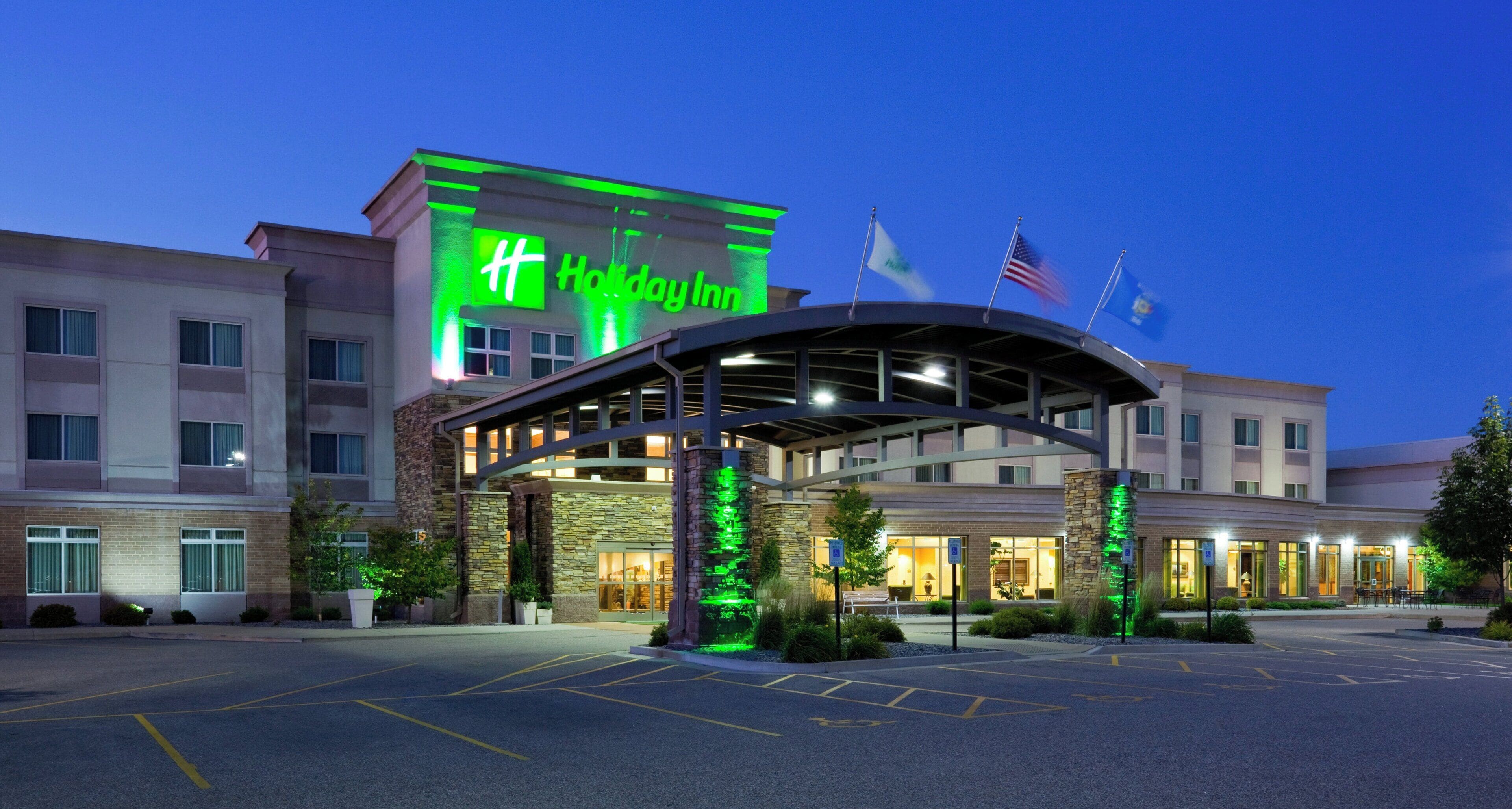 Un hotel Holiday Inn con un diseño moderno, que destaca un letrero verde prominente, un acceso cubierto y varias banderas.