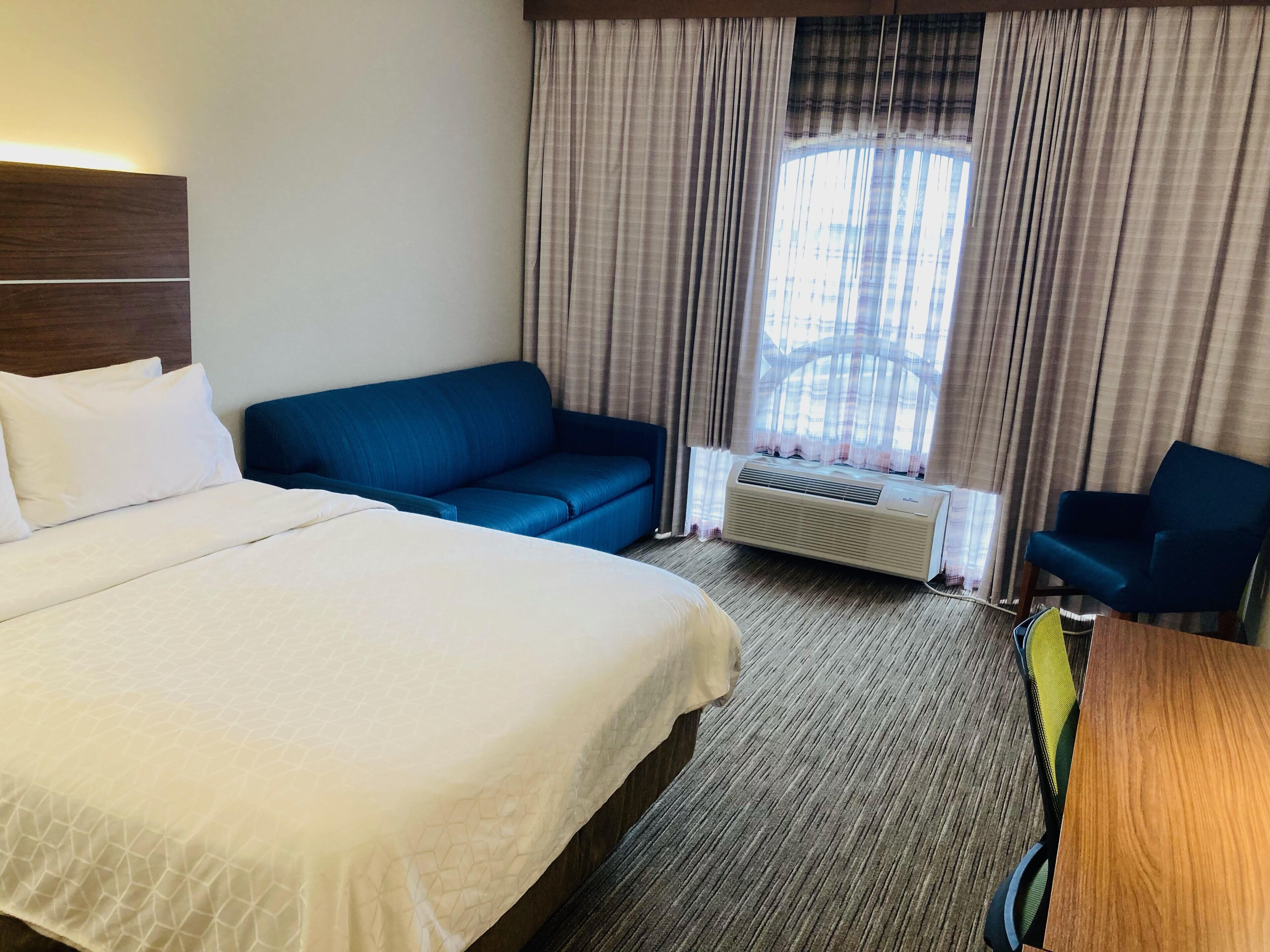 Ett hotellrum med en stor säng, en blå soffa, ett träbord och ett fönster med gardiner.