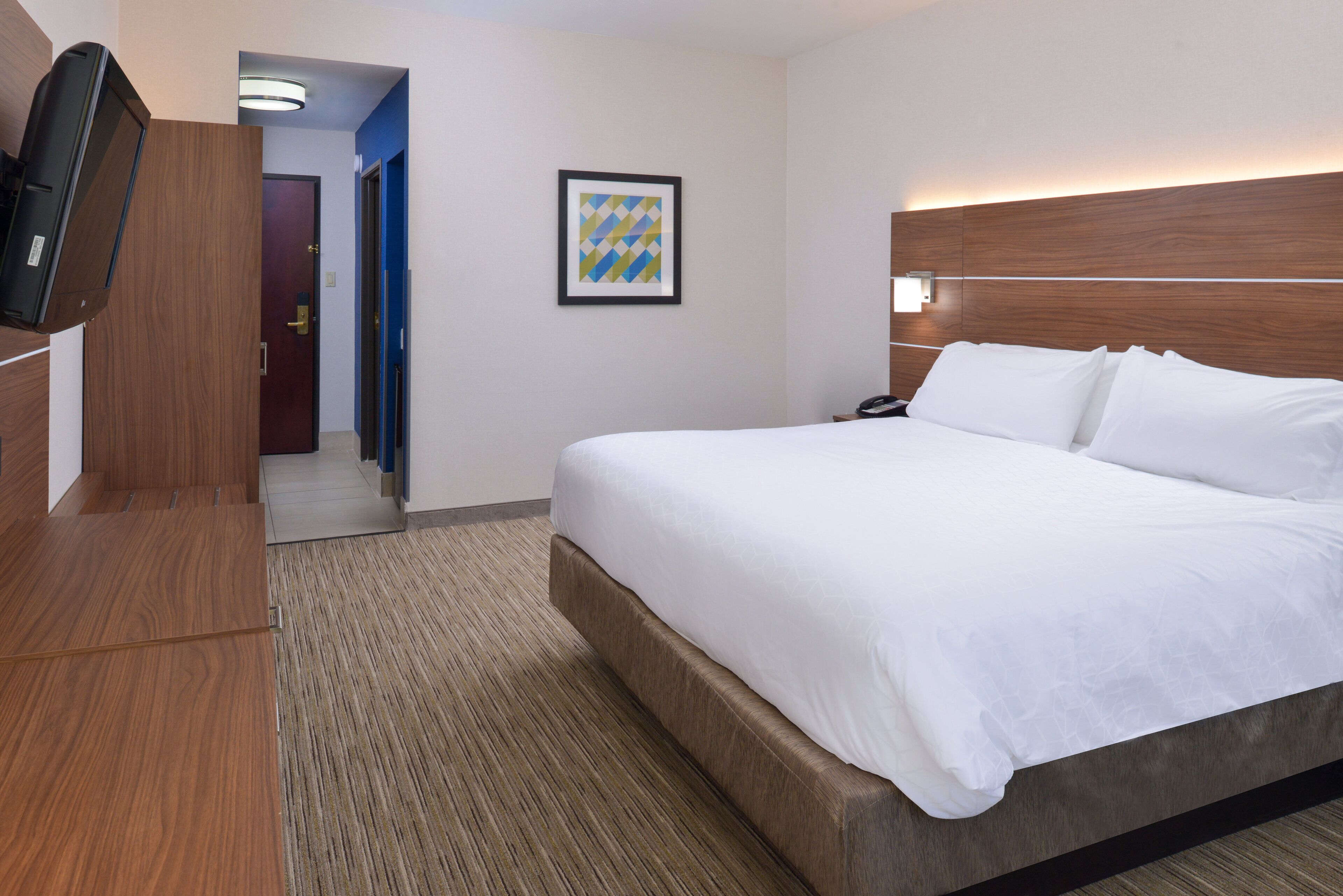 Une chambre d’hôtel avec un grand lit, une tête de lit en bois, un téléviseur à écran plat fixé au mur et une œuvre d’art encadrée accrochée au mur.