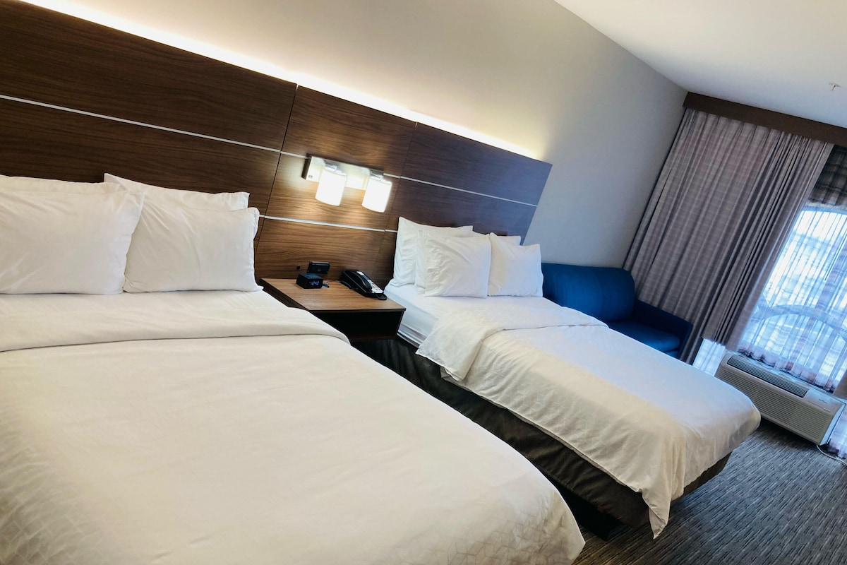 A hotel room with two beds, a nightstand with a phone and a lamp, and a window with curtains.