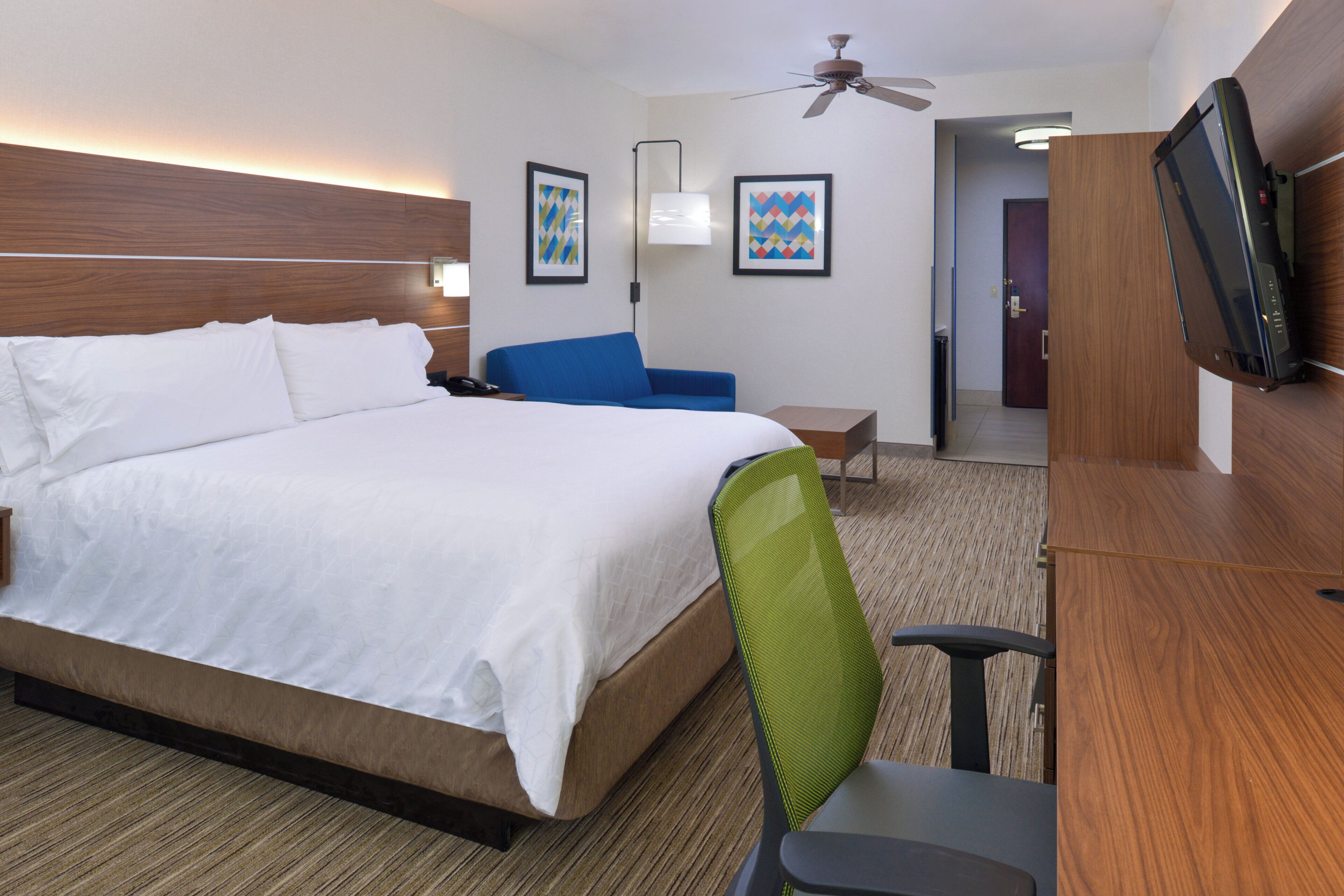 A hotel room with a large bed, a desk, a chair, a television, and two framed artworks on the wall.