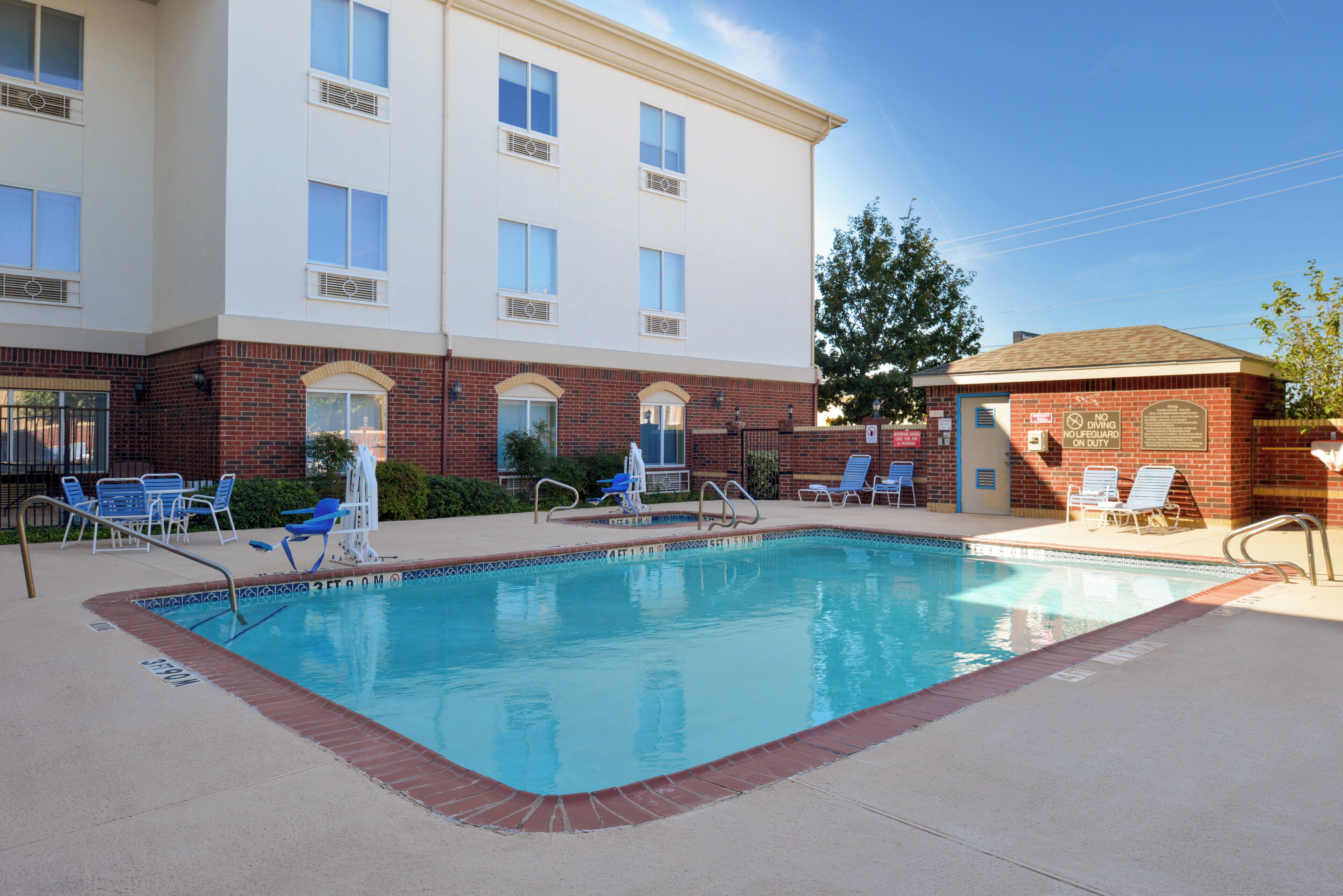 A hotel pool area with a clear blue pool, lounge chairs, and a brick building with multiple windows.