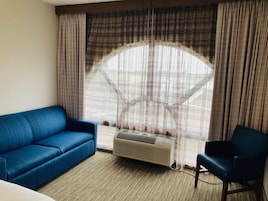 Una habitación con un sofá azul, una silla, un ventanal con cortinas y un equipo de aire acondicionado.