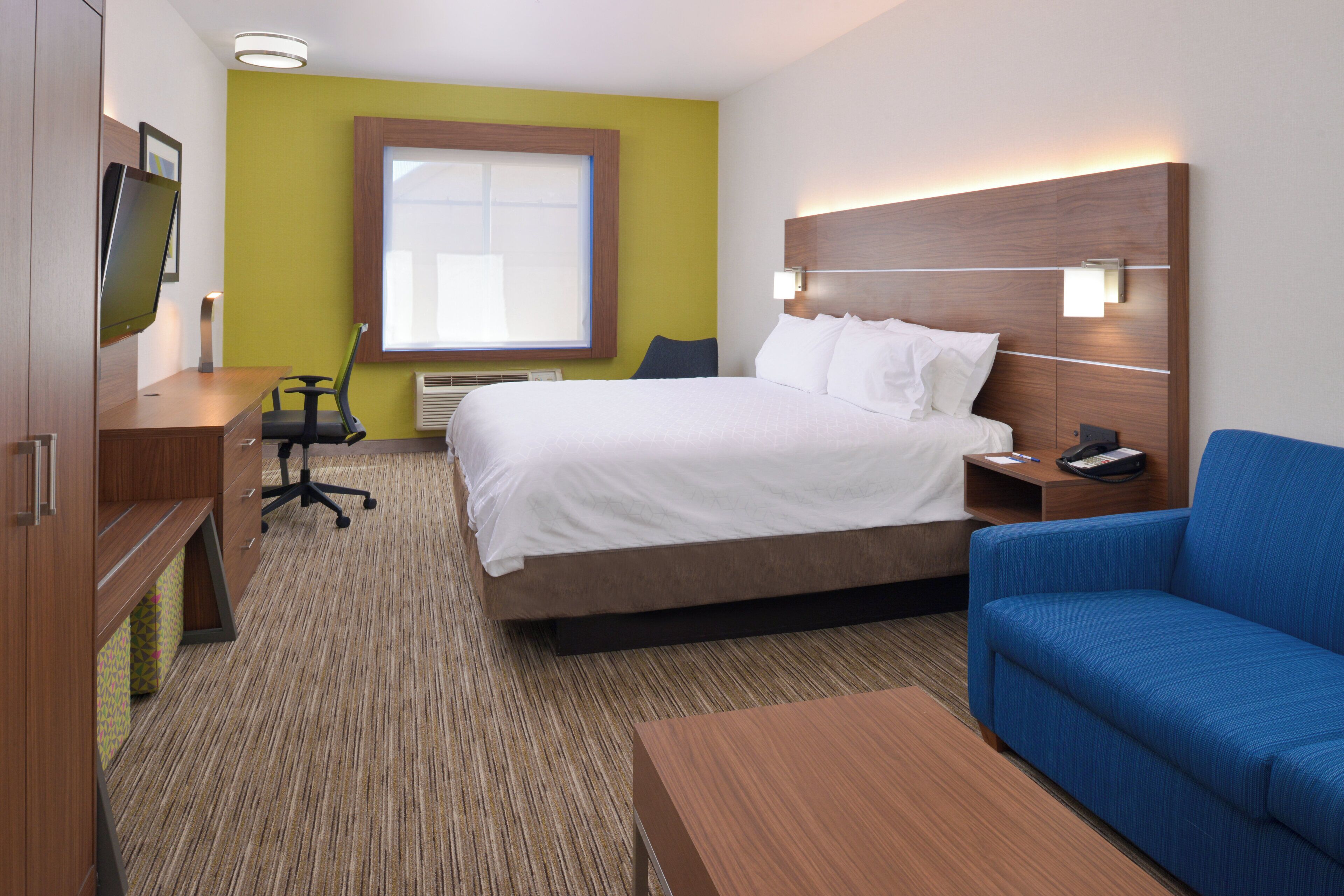 A hotel room with a bed, a desk, a chair, a blue sofa, and a window.