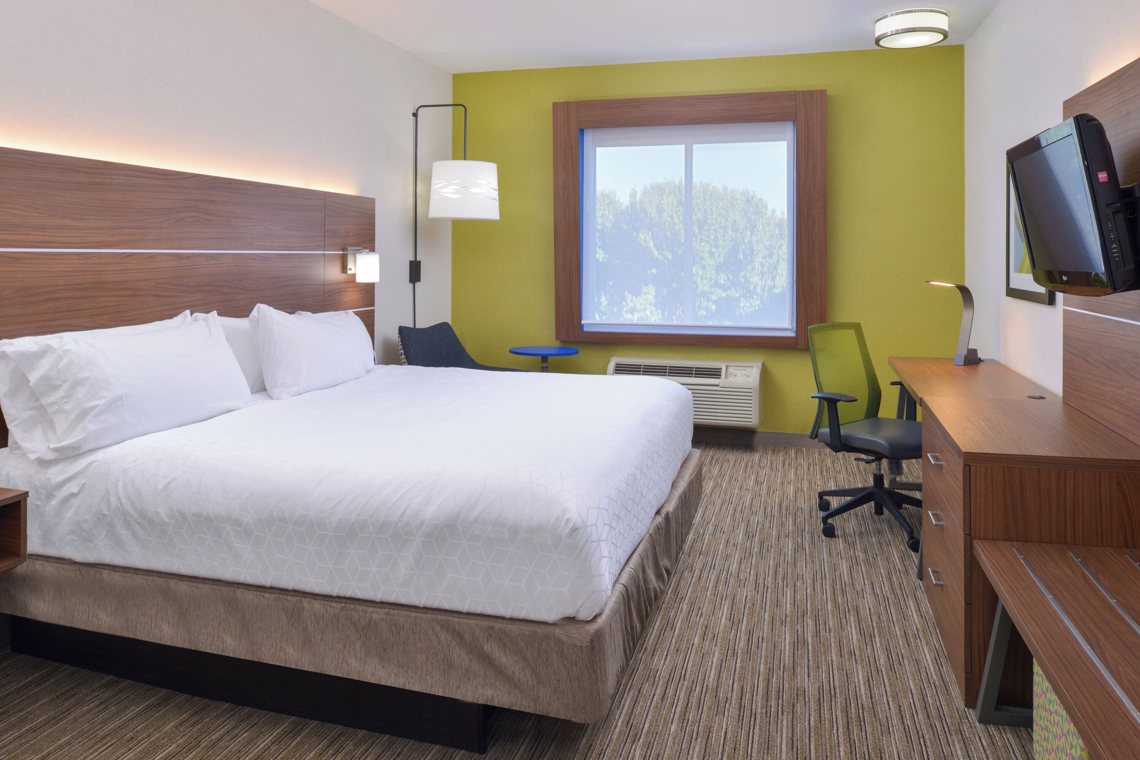 A hotel room with a large bed, a desk, a chair, a television, and a window with a view of trees.