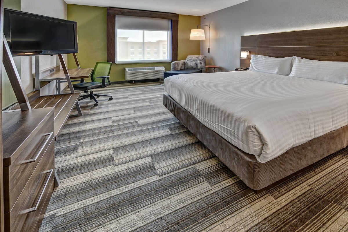 A hotel room with a large bed, a desk with a flat-screen TV, a chair, a small table, and a window with blinds.