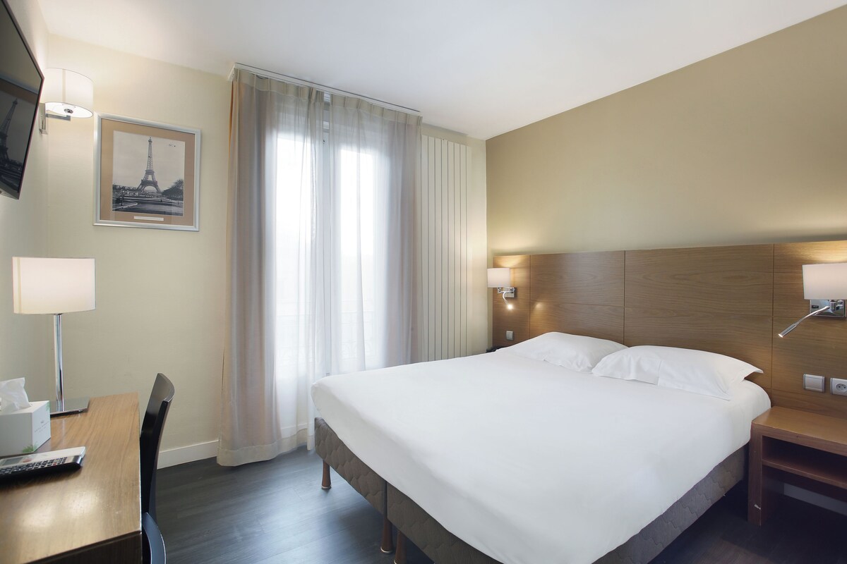 Una habitación de hotel con una cama, mesitas de noche, un escritorio con lámpara, una silla y un ventanal con cortinas.