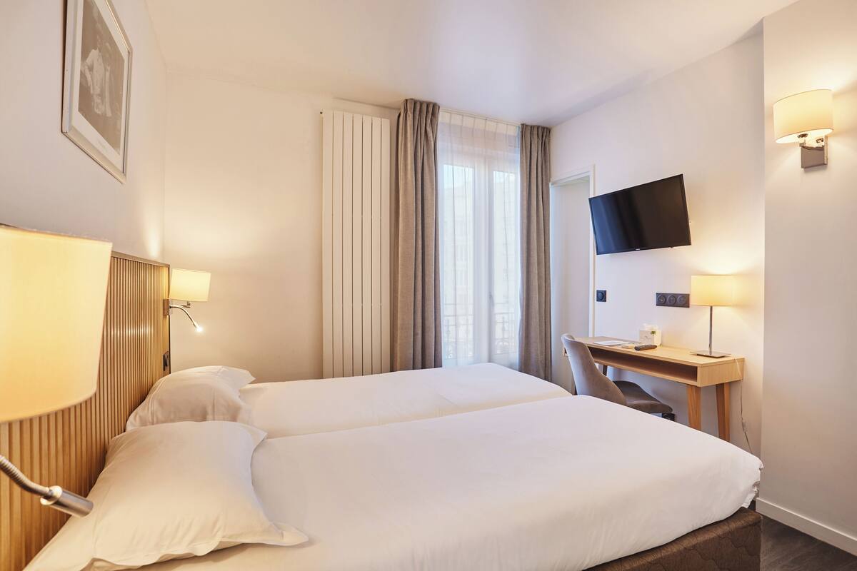 A hotel room with two beds, a desk, a TV, and a window with curtains.