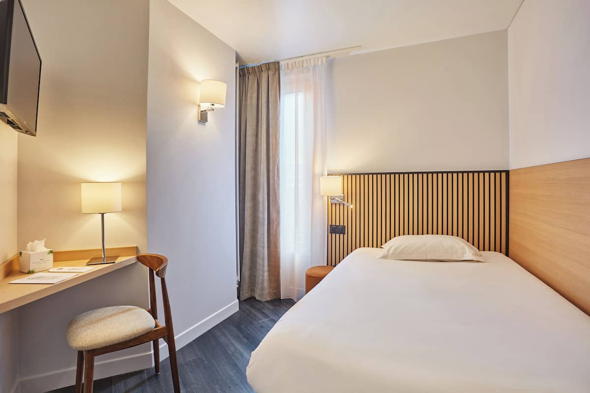 A hotel room with a bed, a desk with a chair, a TV mounted on the wall, and a window with curtains.