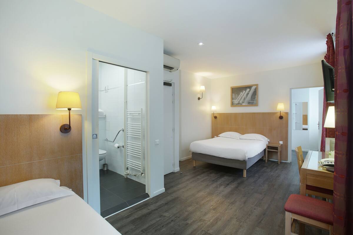 A hotel room with a bed, a small table, a chair, a mirror, a painting, and a bathroom with a shower and toilet.