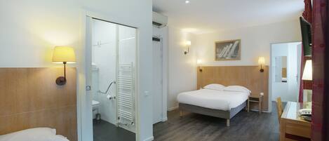 A hotel room with a bed, a small table, a chair, a mirror, a painting, and a bathroom with a shower and toilet.