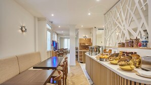 A modern dining area with a long wooden table and chairs, a counter with food items, and a decorative white wall panel. - Hôtel Gabriel Issy (Issy-les-Moulineaux)