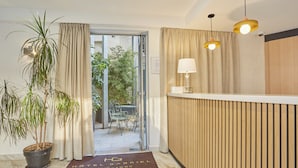 A hotel reception area with a wooden reception desk, a potted plant, and a door leading to a balcony with outdoor seating. - Hôtel Gabriel Issy (Issy-les-Moulineaux)