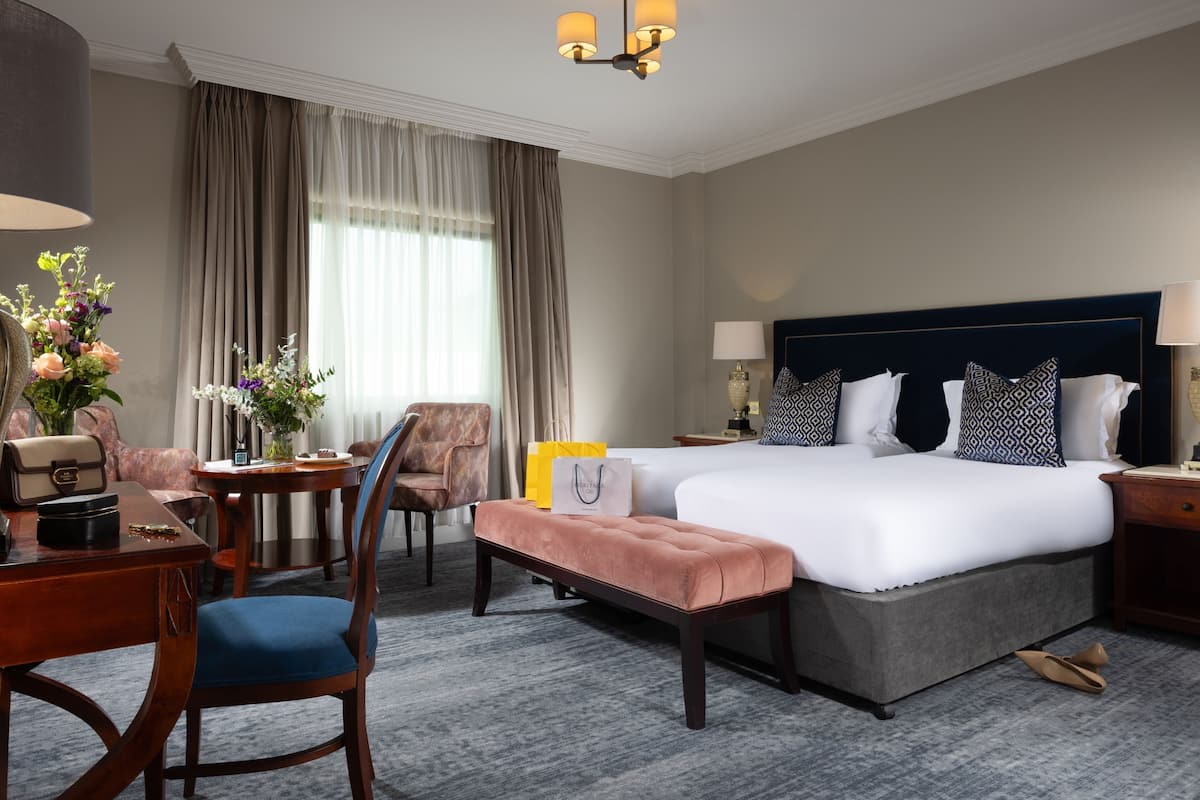 A hotel room with a large bed, a desk with a chair, a bench, a table with a vase of flowers, and a window with curtains.