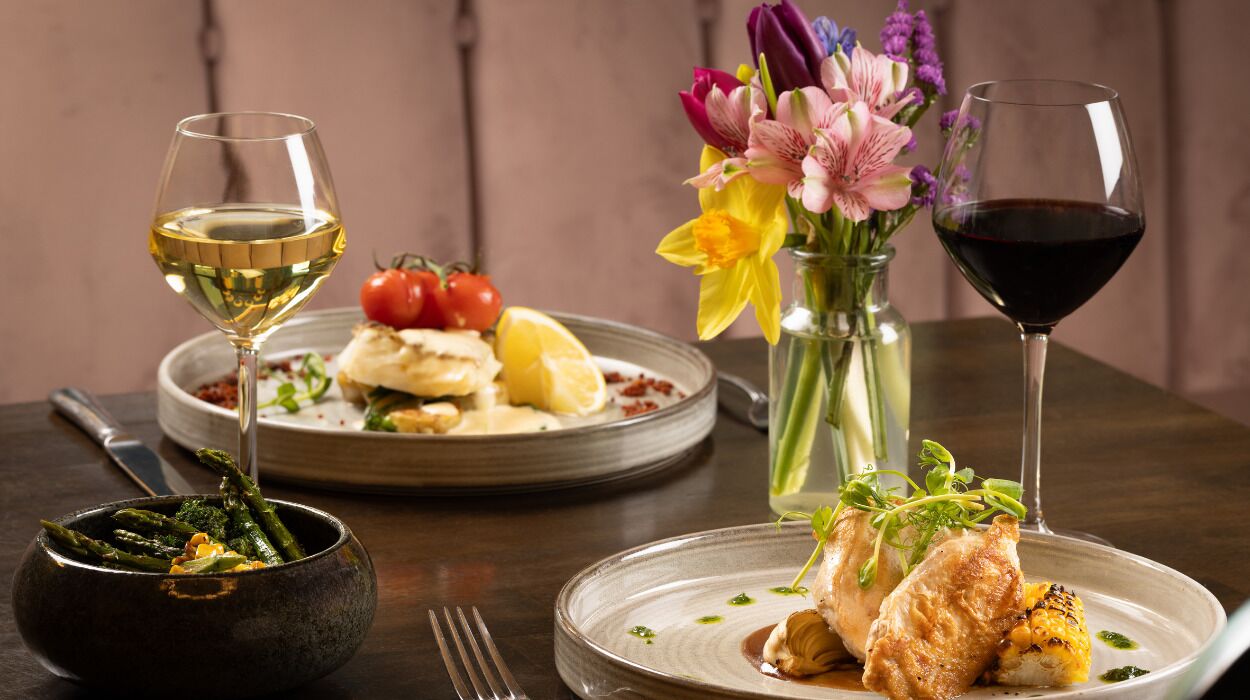 Una mesa de comedor con platos de comida, un bol de espárragos, un jarrón de flores y dos copas de vino.