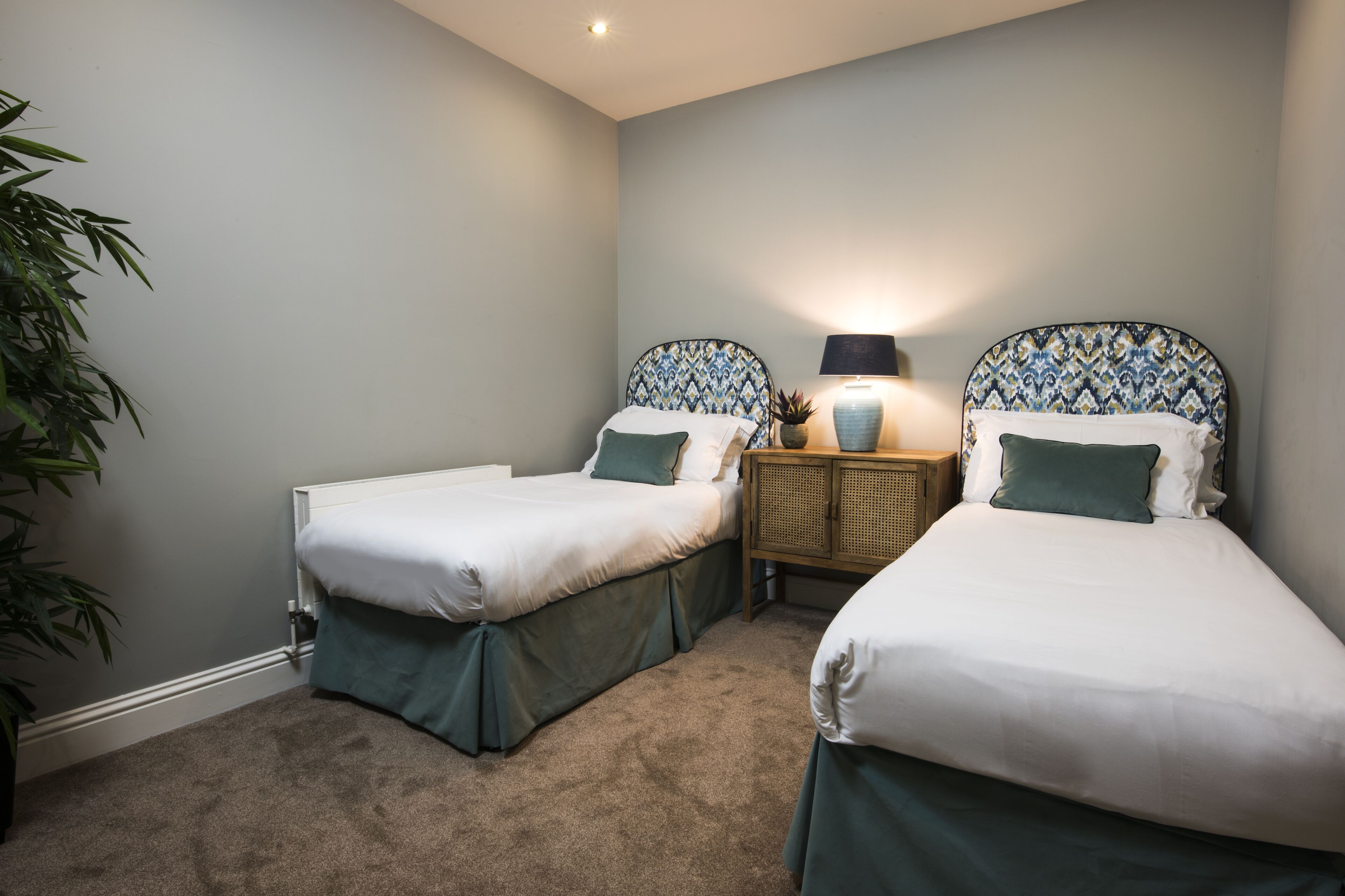 A hotel room with two single beds, a nightstand with a lamp, and a potted plant.