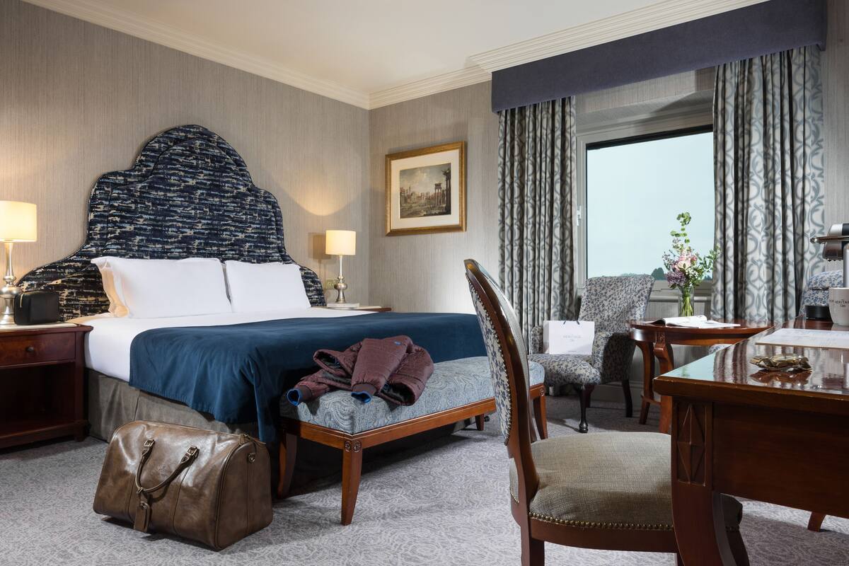 A hotel room with a large bed, a wooden nightstand, a desk, a chair, and a window with curtains.
