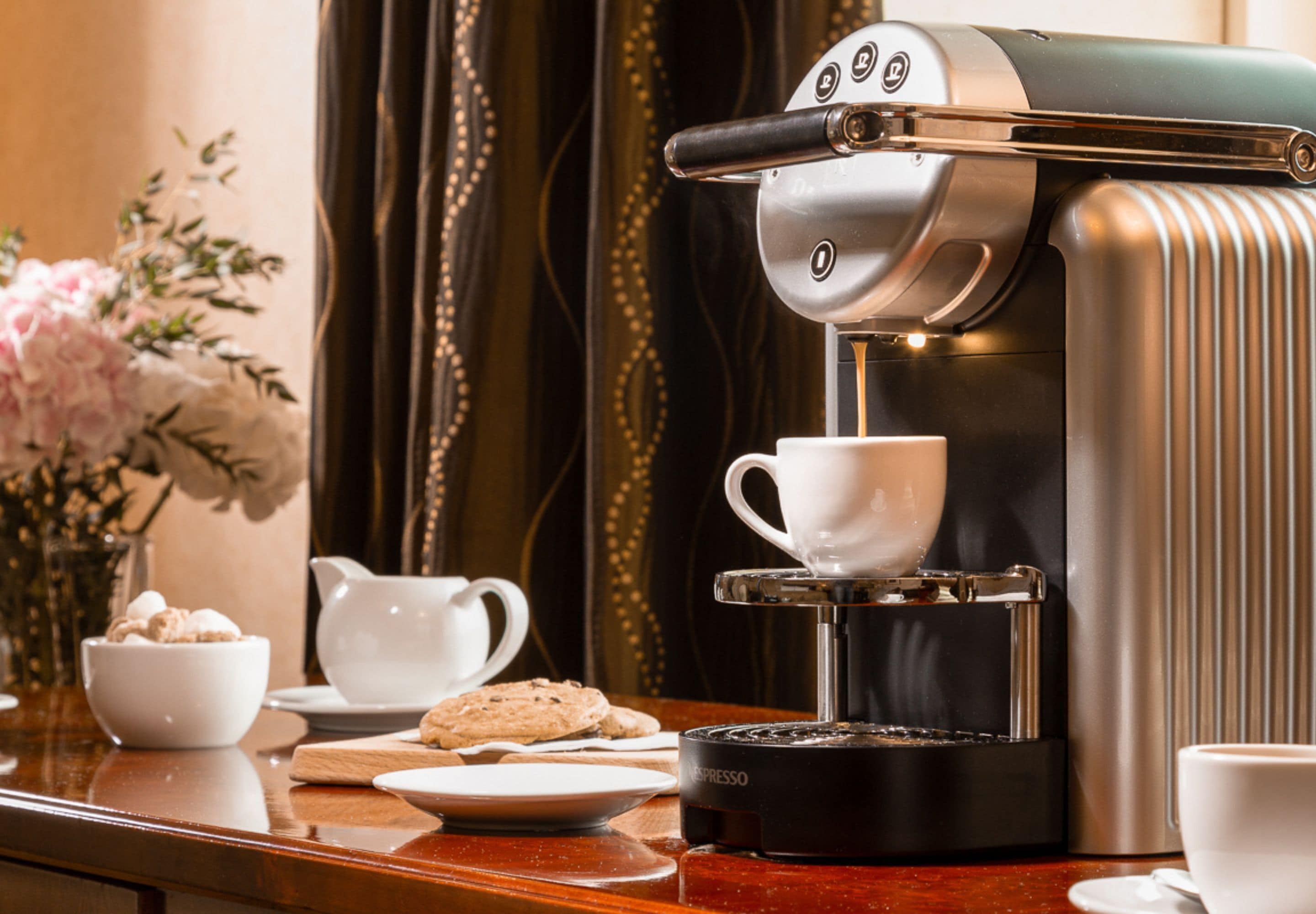 A coffee machine with a cup of coffee being dispensed, a bowl of cookies, and a vase of flowers on a wooden table.