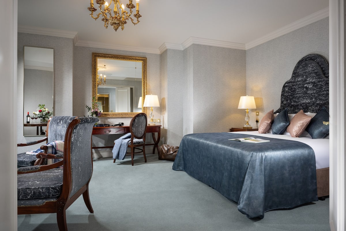 A hotel room with a large bed, a desk with a chair, a dresser, a mirror, and a chandelier.