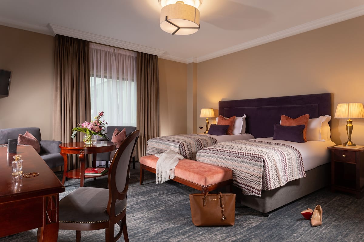 A hotel room with a bed, two bedside tables, a desk, a chair, a small table with a vase of flowers, and a handbag on the floor.