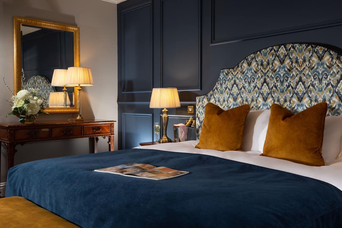 A neatly made bed with a blue bedspread, two lamps on the nightstand, a mirror with a gold frame, and a patterned headboard.